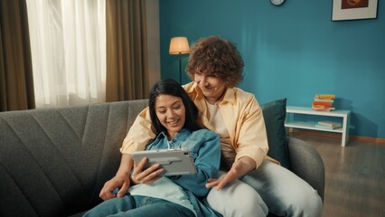 A couple spends time together on a day off. a Woman and a man watch a movie, photo, video on a tablet. A Asian woman lies on a sofa with a tablet in her hands, leaning her back on a man. Close up.