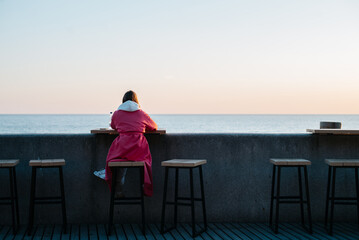 Rear view of a young unrecognizable woman in a pink raincoat resting on the embankment and admiring the seascape