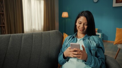 A Asian woman sits on a sofa with a phone in her hands. A woman is typing a message, chatting, scrolling through the feed. Portrait of a smiling woman in the living room on the sofa close up.