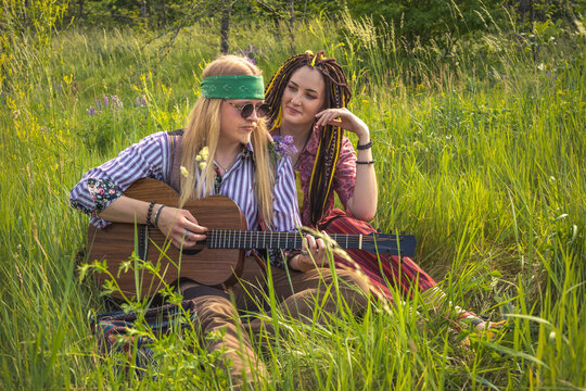Guy and a girl in hippie style with a guitar are sitting in the grass on a forest glade on a sunny day