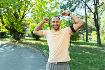 Portrait successful sportsman man satisfied with training results and achievements ,looking at camera and holding hands up in triumph gesture ,hispanic man running in park with headphones and phone.