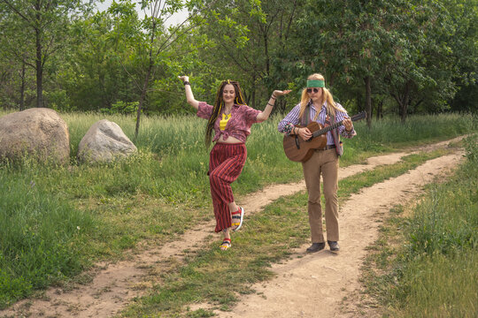 Couple of young people in hippie style. A girl is dancing, a guy is playing a guitar on a country road on a sunny day
