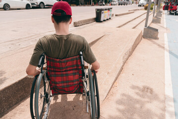Behind of young man with disability pushing wheelchair up ramp on the sidewalk of public road by...