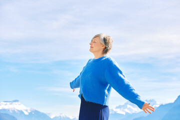 Outdoor portrait of happy middle age 55 - 60 year old woman with arms open wide, posing on blue sky...