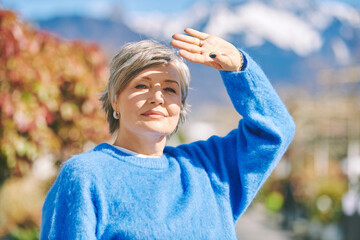 Outdoor portrait of beautiful mature 55 - 60 year old woman, wearing blue pullover, hiding face from the sun with hand - 614413438