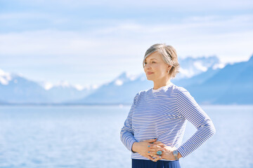 Outdoor portrait of beautiful mature woman posing next to lake, wearing blue stripe t-shirt, healthy lifestyle - 614413488