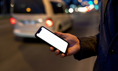 Smartphone with blank screen in hand of passenger on background of night city road traffic, ride-sharing, e-taxis service.