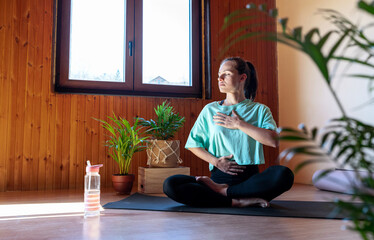 Serene woman sitting on yoga mat at home and doing breathing exercise.