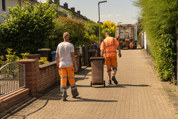 Mörfelden-Walldorf, GERMANY, June 14, 2023 : Garbage workers in orange overalls pull brown garbage cans to a garbage truck for unloading