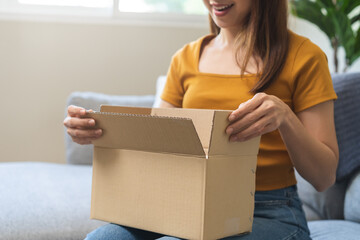 Happy excited, asian young woman, girl customer sitting on sofa at home, open and unpack cardboard box carton parcel after buy fashion clothes, ordering present and shopping online, delivery service.