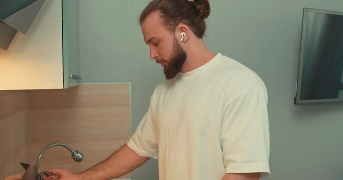 bearded strong young man in white T-shirt with earphones beating eggs at kitchen. close up side view camera moving, slow motion lifestyle free time, household duties, chores