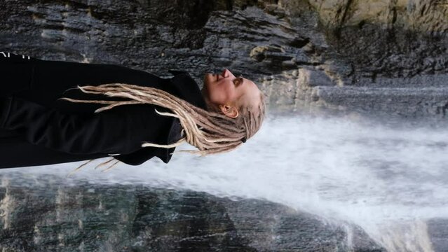 Young woman wearing dreadlocks hairstyle standing near waterfall slow motion vertical video