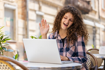 Portrait of pretty young woman using laptop having video call chat conversation for online meeting and presentation outdoors. Girl tourist recording blog sitting in urban sunshine city cafe terrace