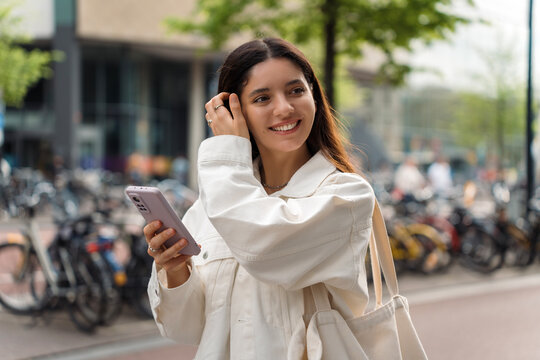 Sustainable young multiethnic woman wearing white eco clothes and a shopper holding a phone walking in the street filled with bicycles in the background