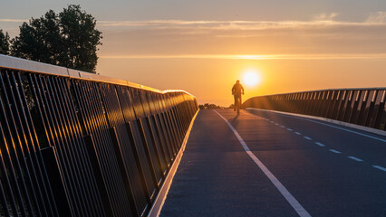 Silhouette of a cyclist crossing a bridge, at sunrise on his way to work - obrazy, fototapety, plakaty