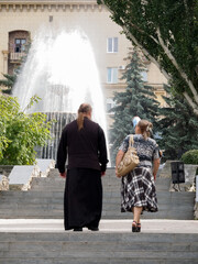 City fountain in the rays of the daytime sun. The priest and the woman climb the steps of the...