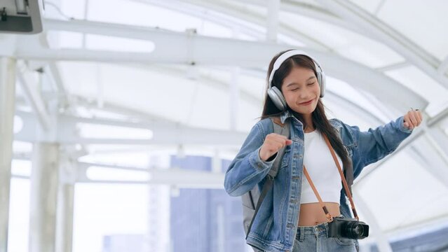 Young asian woman wearing denim jacket, headphones for listening to music and backpack walking in the city. Adolescent female happy, smiling face. Young lady relaxing and enjoying city life