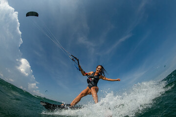 Kitesurfing girl in black sexy swimsuit with kite in sky on board in blue sea riding waves with water splash. Recreational activity, water sports, action, hobby and fun in summer time. 