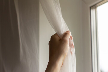 The young man's hands are pulling the tulle curtain on the window.
