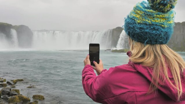 back view of female hiker photographing waterfall with smartphone rainy day,woman traveler taking picture in the wild nature