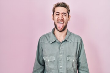 Hispanic man with beard standing over pink background sticking tongue out happy with funny...