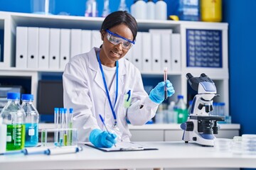 Young african american woman scientist writing on document holding blood test tube at laboratory