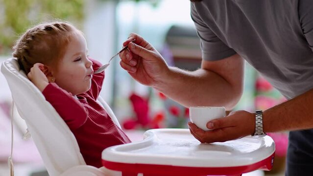 father is feeding cute toddler baby girl in highchair at home