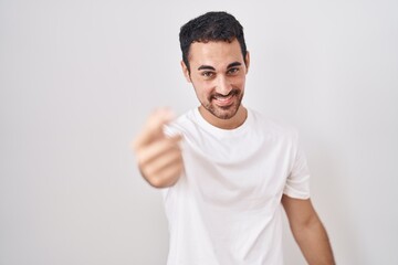 Handsome hispanic man standing over white background beckoning come here gesture with hand inviting welcoming happy and smiling