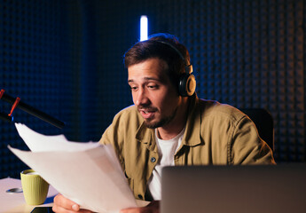 gesturing radio host with headphones reading news from paper into studio microphone at radio...