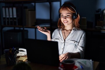 Young caucasian woman working at the office at night smiling cheerful presenting and pointing with palm of hand looking at the camera.