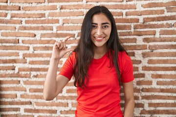 Young teenager girl standing over bricks wall smiling and confident gesturing with hand doing small size sign with fingers looking and the camera. measure concept.