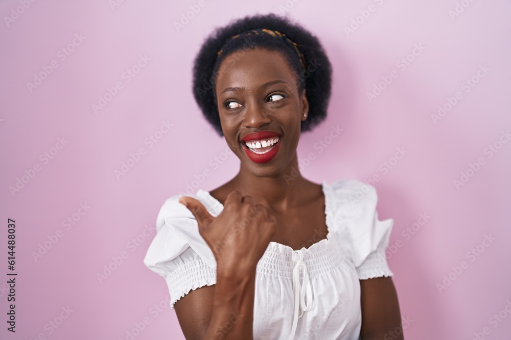 Wall mural African woman with curly hair standing over pink background smiling with happy face looking and pointing to the side with thumb up.