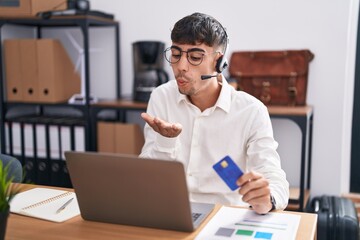 Young hispanic man working using computer laptop holding credit card looking at the camera blowing a kiss with hand on air being lovely and sexy. love expression.