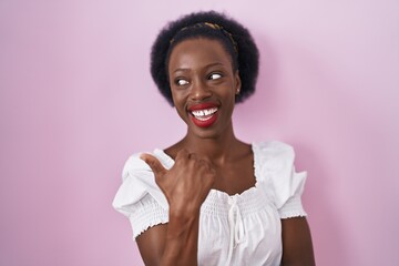 African woman with curly hair standing over pink background smiling with happy face looking and pointing to the side with thumb up.