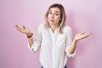 Young beautiful woman standing over pink background clueless and confused expression with arms and...