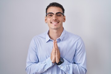 Handsome hispanic man wearing business clothes and glasses praying with hands together asking for forgiveness smiling confident.