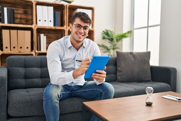 Young hispanic man psychologist smiling confident using touchpad at psychology clinic