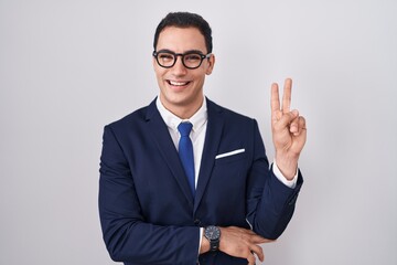 Young hispanic man wearing suit and tie smiling with happy face winking at the camera doing victory sign. number two.