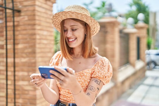 Young redhead woman tourist wearing summer hat watching video on smartphone at street