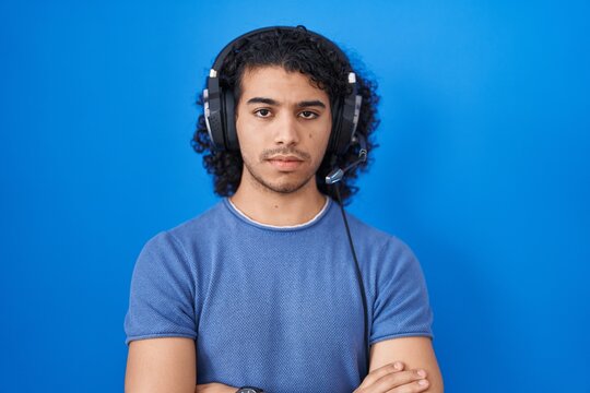 Hispanic man with curly hair listening to music using headphones skeptic and nervous, disapproving expression on face with crossed arms. negative person.