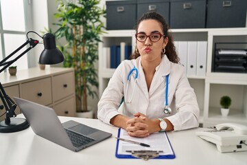 Young hispanic woman wearing doctor uniform and stethoscope puffing cheeks with funny face. mouth inflated with air, crazy expression.