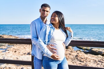 Young latin couple expecting baby hugging each other standing at seaside