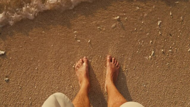 POV male feet standing on golden sand with sea water foam. First person view of man on ocean beach at golden hour. Slow motion view of waves rolling on feet and brown sand. Idyllic summer holiday.