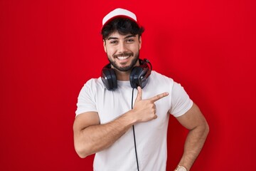 Hispanic man with beard wearing gamer hat and headphones cheerful with a smile on face pointing with hand and finger up to the side with happy and natural expression