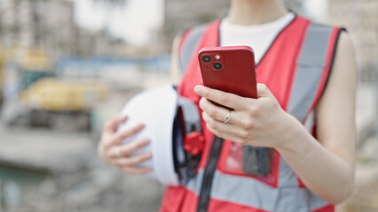 Young beautiful hispanic woman builder using smartphone holding hardhat at street