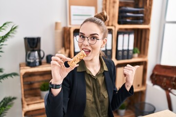 Young hispanic girl eating protein bar as healthy energy snack at the office pointing thumb up to the side smiling happy with open mouth