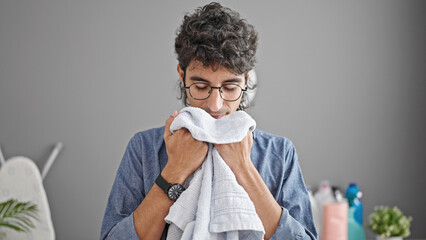 Young hispanic man smiling confident smelling clean towel at laundry room