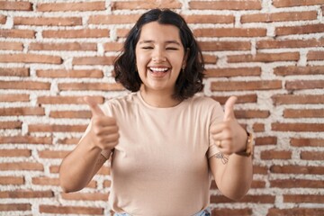 Young hispanic woman standing over bricks wall success sign doing positive gesture with hand,...