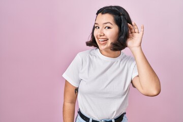 Young hispanic woman wearing casual white t shirt over pink background smiling with hand over ear listening an hearing to rumor or gossip. deafness concept.