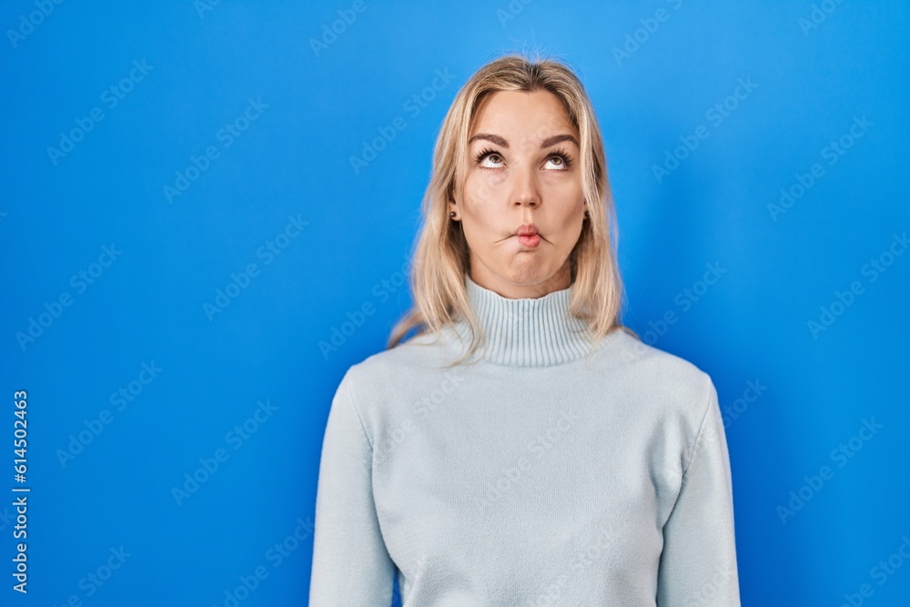 Sticker Young caucasian woman standing over blue background making fish face with lips, crazy and comical gesture. funny expression.